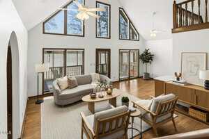 A living room with high vaulted ceilings, large triangular and rectangular windows, two ceiling fans, a gray sofa, two armchairs, a round coffee table, a sideboard, houseplants, and modern decor.