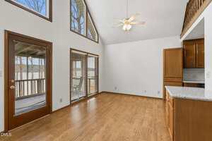 A bright, empty room with wood floors, large windows, a ceiling fan, a glass door to a balcony, and wood cabinets with granite countertops on the right. Loft railing visible above the kitchen area.