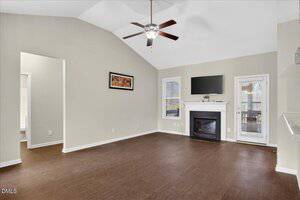 A spacious living room with beige walls, wood flooring, a white fireplace under a wall-mounted TV, and two windows with blinds. There is a ceiling fan and a framed artwork on one wall.