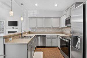 A modern kitchen with gray cabinets, stainless steel appliances, a double sink, a light-colored countertop, tile backsplash, and wood flooring. Pendant lights hang above the counter, and an open living area is partially visible.