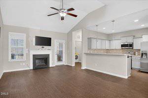 A modern open-concept living area with wood floors, a ceiling fan, wall-mounted TV above a fireplace, and a kitchen with white cabinets, stainless steel appliances, and pendant lights. A laundry area is visible through a doorway.