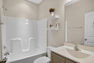 A bathroom with a white bathtub and shower, beige walls, a toilet, a sink with a beige countertop, a wall mirror, towel rack, and two white shelves holding decorative items above the toilet.