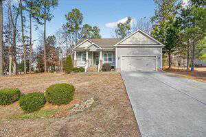 A single-story beige house at 15 Saddletree Drive, Franklinton, features a front porch, attached two-car garage, and concrete driveway. The yard has sparse grass, some shrubs, and is surrounded by tall trees under a clear sky.