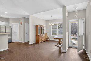 A dining area with wood flooring features a round table, a wooden hutch, and two chairs near windows. An open door leads outside, and part of a kitchen with white cabinets and a stainless steel fridge is visible.