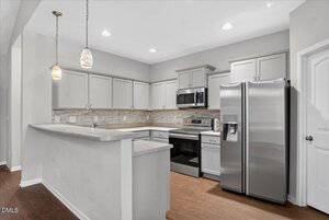 Modern kitchen with light gray cabinets, stainless steel appliances, a double-door refrigerator, built-in microwave, electric stove, light-colored countertops, backsplash, and two pendant lights over a breakfast bar.