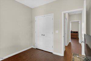 A room with beige walls, a white double-door closet, wood flooring, and a view into a hallway with a dresser visible in another room. A small piece of furniture is partially seen in the lower right corner.