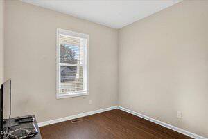 A small, empty room with beige walls, a window with white blinds, dark wood flooring, and a TV on a stand in the lower left corner. Sunlight comes in through the window.
