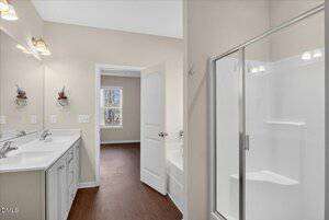 A bathroom with a double sink vanity, a glass shower, a bathtub, beige walls, and wood flooring. An open door leads to a bright room with a window and similar flooring.