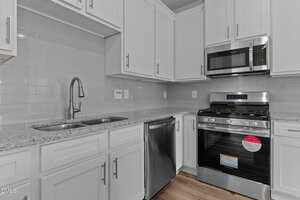A modern kitchen with white cabinets, granite countertops, a stainless steel double sink, dishwasher, gas stove with oven, and microwave. The backsplash is white subway tile and the floor is light wood.