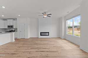 A spacious, empty living room with light wood floors, white walls, a ceiling fan, large window, and a built-in electric fireplace. The open layout connects to a white kitchen with stainless steel appliances.