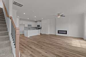 Open-plan living area with light wood flooring, white walls, a ceiling fan, an electric fireplace, and an adjacent kitchen featuring white cabinetry and a kitchen island. Carpeted stairs with a wooden handrail are on the left.