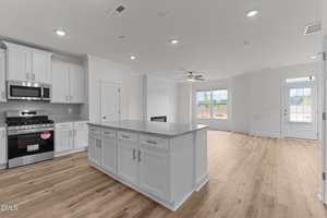 A modern open-concept kitchen with white cabinets, stainless steel appliances, a central island, and light wood flooring. The kitchen opens to a spacious living area with large windows and a ceiling fan.