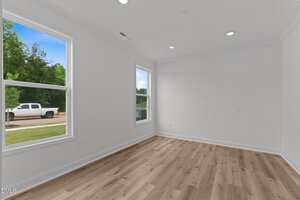 An empty room with white walls, recessed ceiling lights, two large windows letting in natural light, and light-colored wood flooring. A white truck and greenery are visible outside through the windows.