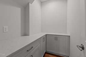 A small, modern room with white walls, gray cabinets, and a white marble countertop. There is a visible electrical outlet on the left wall and a wooden floor. The space is clean and empty.