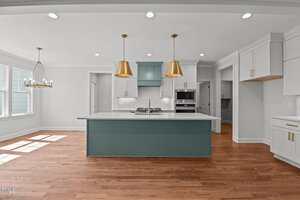 A modern kitchen with wood floors, white cabinets, a green island, gold pendant lights, a white countertop, stainless steel appliances, and a large window bringing in natural light.