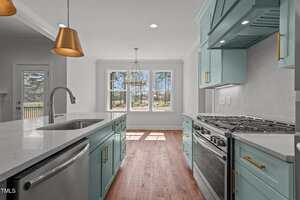 A modern kitchen features teal cabinets, stainless steel appliances, gold hardware, pendant lights, and a large window overlooking a yard. White countertops and backsplash create a clean, bright atmosphere.