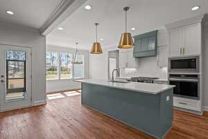 A modern kitchen with hardwood floors, a large blue-gray island with white countertops, two gold pendant lights, white cabinets, built-in oven and microwave, and a windowed breakfast nook.