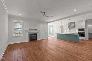 Modern open-concept living area with hardwood floors, a white fireplace, ceiling fan, large windows, and an adjacent kitchen featuring a teal island, gold pendant lights, and white cabinetry.