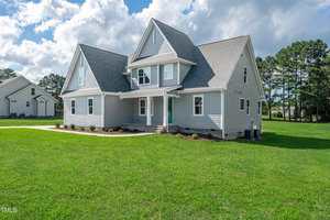 A two-story gray house with white trim and a covered front porch stands on a large, well-kept lawn. The house has multiple gables, an attached garage, and is surrounded by trees and a clear sky with clouds.