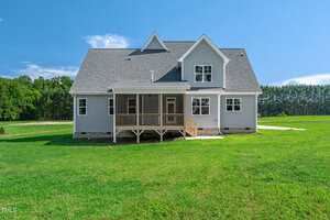 A gray two-story house with white trim, a covered front porch, and multiple windows, set on a grassy lawn with trees in the background under a clear blue sky.