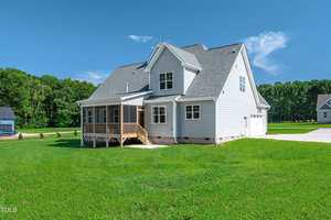 A two-story white house with gray roofing sits on a large, neatly mowed green lawn. The home features a screened porch, multiple windows, and an attached two-car garage. Trees line the background.