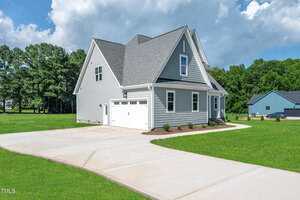 A modern gray house with white trim and a two-car garage sits on a large, well-maintained lawn. The driveway is wide and concrete, and there are trees and another house in the background under a partly cloudy sky.