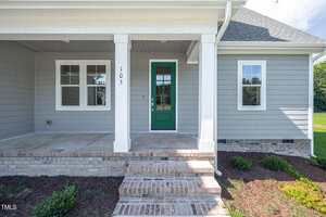 The image shows the front entrance of a modern house with light gray siding, a green door with glass panels, white trim, brick steps, and a small covered porch. The house number 105 is visible on a column.
