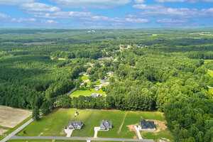 Aerial view of a rural neighborhood with three houses in the foreground, surrounded by green fields and dense wooded areas under a partly cloudy sky. More homes and trees are visible in the background.