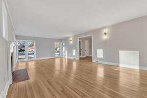 Spacious, empty living room with light wood floors, light-colored walls, and several windows letting in natural light. There is a fireplace on the left and built-in shelves in an alcove at the back.
