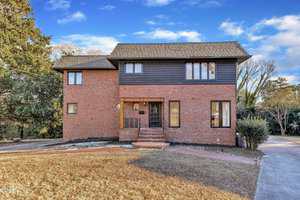 This two-story brick house at 126 Person Street, Louisburg, features dark trim, a central entrance, and multiple rectangular windows. Set on a dry brown lawn with a concrete driveway, it has trees in the background beneath a blue sky with clouds.