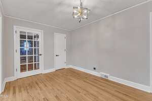 A bright, empty room with light gray walls, hardwood flooring, a modern geometric ceiling light fixture, a white door, and a double glass door. A floor vent and electrical outlet are visible on one wall.