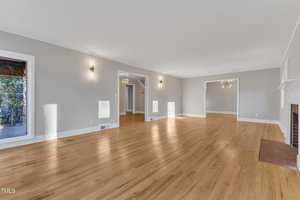 A spacious, empty living room with light gray walls, hardwood floors, wall-mounted lights, large windows, and a brick fireplace. Open doorways lead to adjoining rooms, including a dining area with a chandelier.