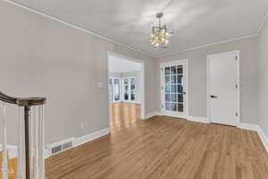 A room with light hardwood floors, beige walls, white trim, a modern ceiling light fixture, and three doorways: one with glass double doors, one single door, and an open entryway leading to a sunlit room.