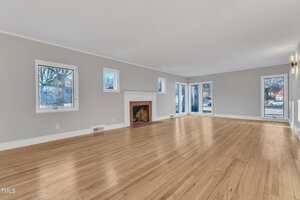 Spacious, empty living room with light-colored wooden floors, light gray walls, white trim, a white fireplace with a red brick interior, and multiple windows letting in natural light.