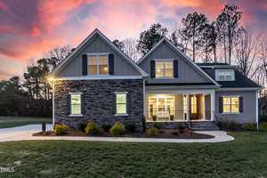 Modern two-story house at 115 Old Garden Lane, Youngsville, featuring stone and gray siding exterior, large windows, front porch with white columns, and a well-maintained lawn, photographed at sunset with trees in the background.
