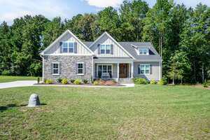 A modern two-story house with light gray siding, stone accents, and a covered front porch, surrounded by a well-kept lawn and trees in the background under a clear sky.