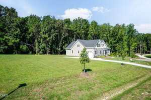 A two-story house with stone and siding exterior sits on a large, grassy lot bordered by trees. A driveway leads to the house, and there is a small tree planted in the front yard. The sky is partly cloudy.