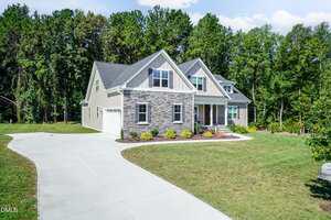 A two-story suburban house with gray siding, stone accents, and a gabled roof. The property has a large driveway, landscaped front yard, and is surrounded by trees and greenery under a clear sky.