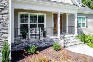 A front porch of a house with gray siding and stone accent, black metal railing, a wooden front door with glass panels, two potted plants, and a white bench with decorative pillows near a large window.