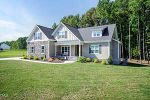 A modern single-story house with gray siding, stone accents, and a covered front porch sits on a large grassy yard with landscaped shrubs and trees in the background under a clear blue sky.