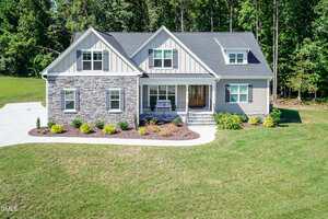 A two-story suburban house with gray siding and stone accents, a front porch with two chairs, manicured landscaping, and a large grassy lawn bordered by trees in the background.