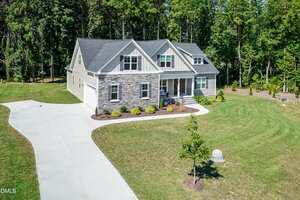 A two-story suburban house with stone and siding exterior, a covered front porch, and a large curved driveway set on a spacious, grassy lot with trees in the background.