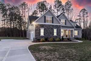 A modern, two-story house with gray siding and stone accents, large windows, and a front porch. The driveway curves toward the attached garage, and the lawn is bordered by trees at sunset.