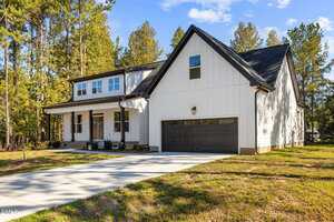 A modern two-story house with white siding, black trim, a front porch, and a double garage sits on a concrete driveway surrounded by grass and tall trees under a blue sky.