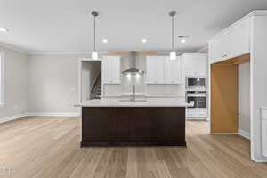 Modern kitchen with light wood flooring, white cabinets, stainless steel appliances, a large dark island with sink, two pendant lights above, and an open pantry or fridge space on the right.