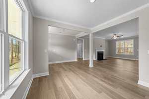 A spacious, empty living and dining area with light wood floors, neutral-colored walls, large windows, a ceiling fan, and a fireplace, with natural light streaming in from outside.