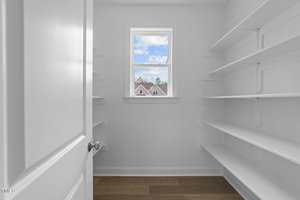 A small, white walk-in pantry with open shelves on both sides, a wooden floor, and a window at the far end showing a view of houses and trees under a blue sky with clouds.