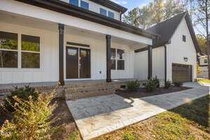 A modern white house with dark trim, a covered front porch, brown double front doors, brick steps, landscaped shrubs, and a paved walkway leading up to the entrance.