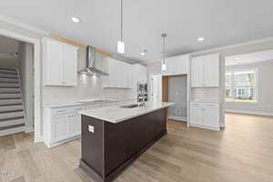Modern kitchen with white cabinets, stainless steel appliances, a dark central island with a sink, light wood flooring, recessed lighting, and a window providing natural light. Stairs are visible to the left.