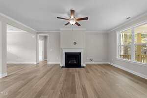 A spacious, empty living room with light wood floors, beige walls, a white ceiling, a ceiling fan with lights, a white fireplace, and large windows letting in natural light. An open doorway leads to another room.